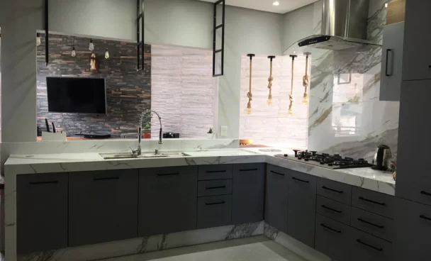 A imagem ilustra uma cozinha americana planejada na cor cinza escuro com uma pedra de mármore branco em cima e detalhes metalizados e pretos, como a pia e a geladeira.