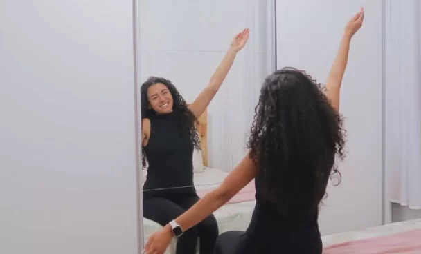 Uma mulher com cabelos cacheados, vestindo roupa preta, sentada na cama de frente para um espelho grande, com os braços levantados e sorrindo.