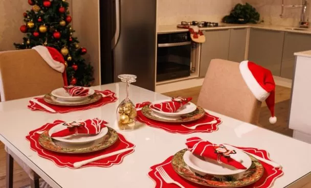 Mesa posta de Natal Madesa, decorada com sousplat vermelho e decorações douradas. Ao fundo, uma cozinha Madesa decorada e uma árvore de Natal.